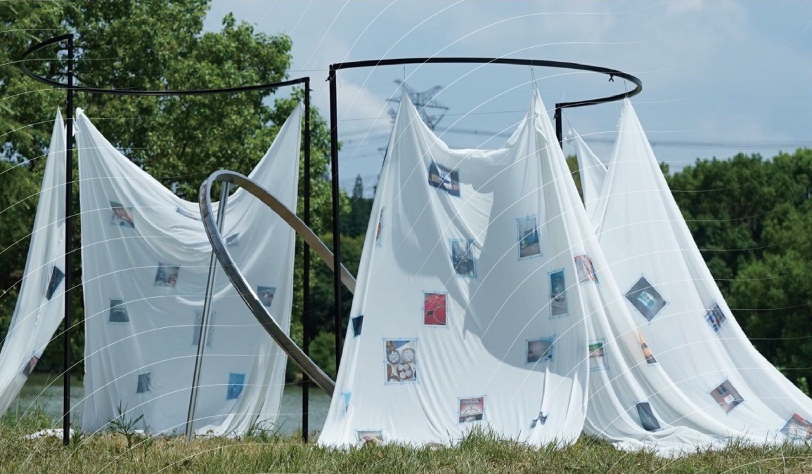 Spiral installation — white fabric draped over steel rings by a river, printed memories pinned across it